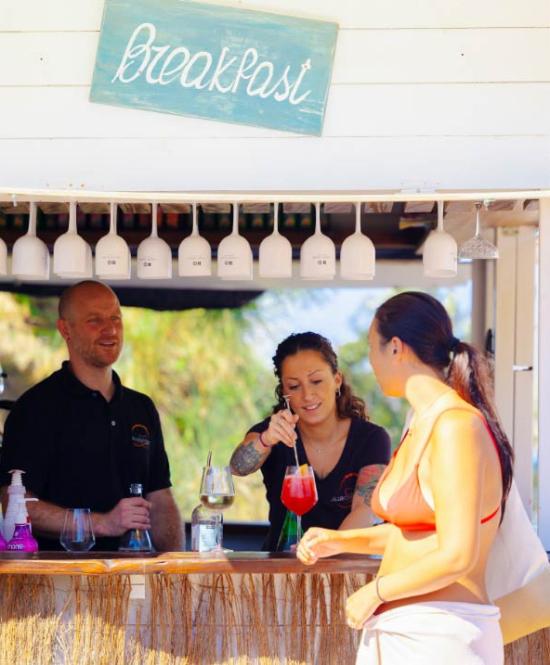 Chiosco colazione sulla spiaggia con persone sorridenti e cocktail.